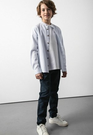 Jasnoniebieska koszula na guziki noszona na białej koszulce, zestawiona z ciemnymi jeansami i białymi tenisówkami. Posiada kieszeń na piersi i drewniane guziki.
