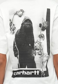 Fehér póló, amelyen fekete-fehér grafika látható: egy alakot szárított rózsák vesznek körül, alul pedig a "carhartt" márkanév.