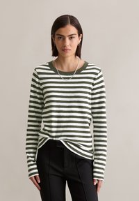 Woman with short dark hair wearing silver chain necklace, green and white striped long-sleeve shirt, and black pants against beige background.
