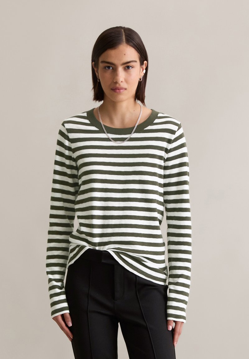 Woman with short dark hair wearing silver chain necklace, green and white striped long-sleeve shirt, and black pants against beige background.