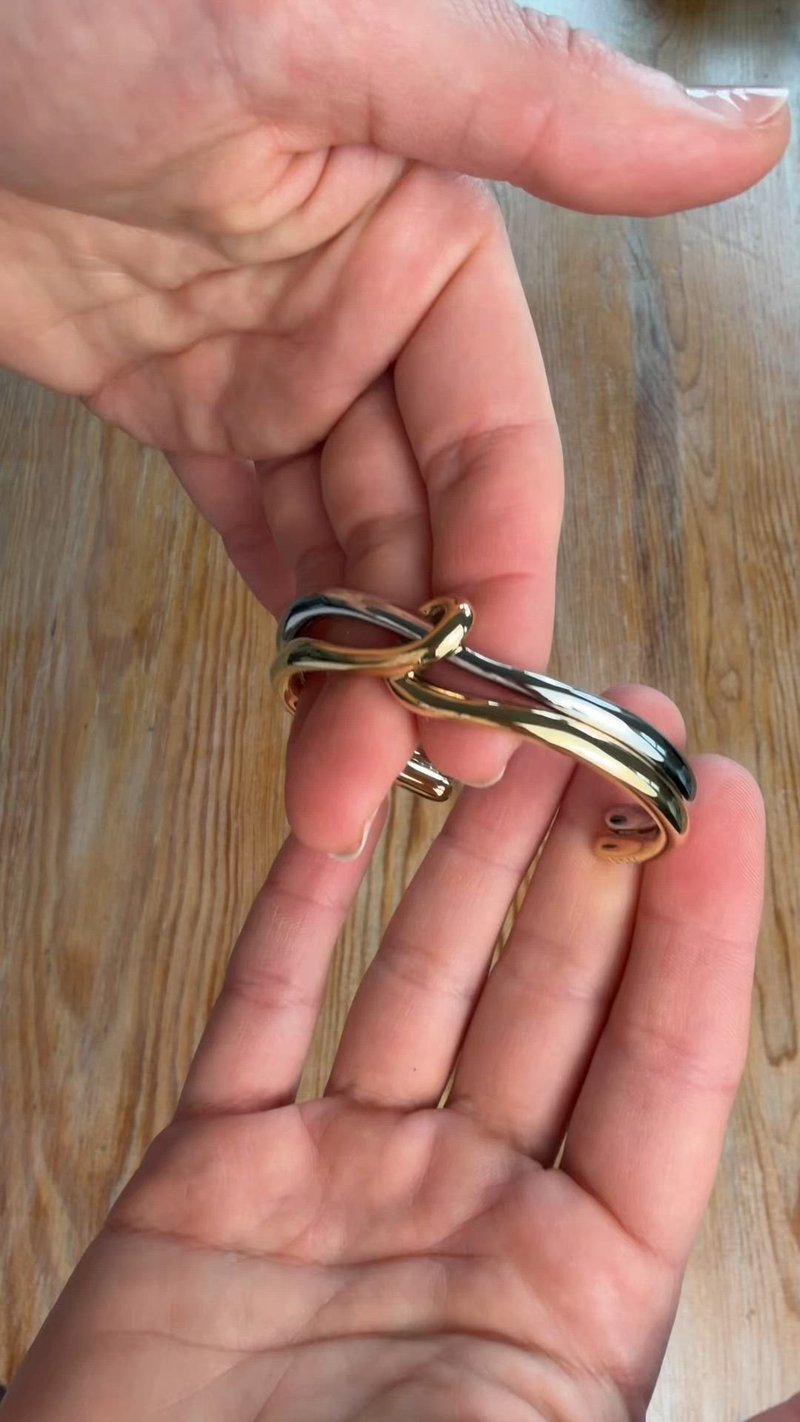 Deux mains tenant un bracelet en métal poli torsadé, présentant des bandes entrelacées en or et argent, au-dessus d'une surface en bois.