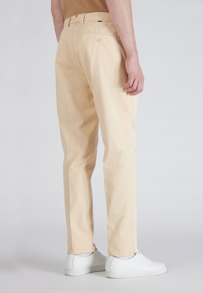 Chinos beige chiaro con una texture liscia, taglio dritto, due tasche posteriori con chiusura a bottone, abbinati a sneakers bianche.