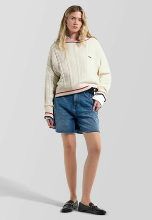 Giovane donna che indossa un maglione crema a trecce con rifiniture rosse e nere, pantaloncini di jeans e mocassini neri, in piedi davanti a uno sfondo grigio.