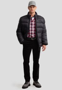Veste rembourrée noire à col montant sur une chemise à carreaux rouge et bleu, accompagnée de pantalons en cord black et de bottes de randonnée grises.