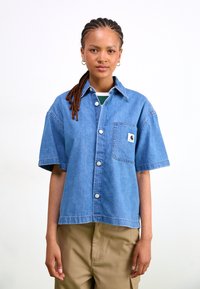 Short-sleeve denim shirt in light blue with a chest pocket, button-down front, and a brand logo patch. Smooth texture and relaxed fit.