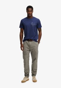 Camiseta de algodón azul marino con un logo estampado en el pecho izquierdo, combinada con pantalones cargo gris claro con bolsillos laterales, usada con zapatillas beige.