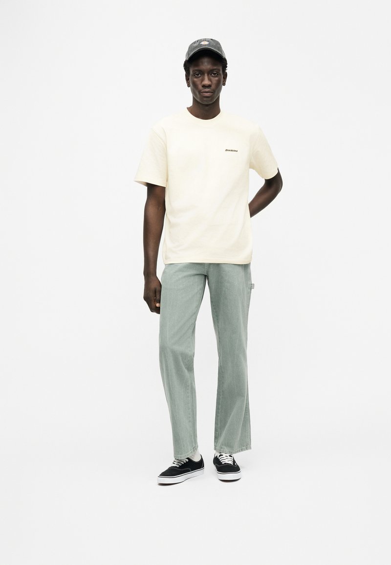 Maglietta crema a maniche corte con logo, pantaloni verde chiaro a gamba larga e sneakers nere. Il modello indossa un cappello grigio e posa su uno sfondo bianco.