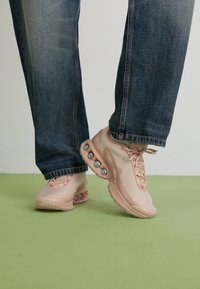 Light pink sneakers with a smooth upper, round accents on the sole, and laces, paired with blue denim jeans on a green surface.