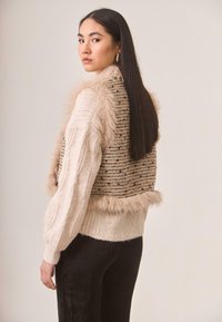 Chaleco de punto beige con cuello de piel y espalda texturizada, que presenta un patrón a rayas con acentos oscuros, combinado con pantalones negros.