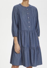 Modré šaty ke kolenům s texturovaným vzorem listů, dlouhé rukávy s elastickými manžetami a předním zapínáním na knoflíky na stojící osobě.