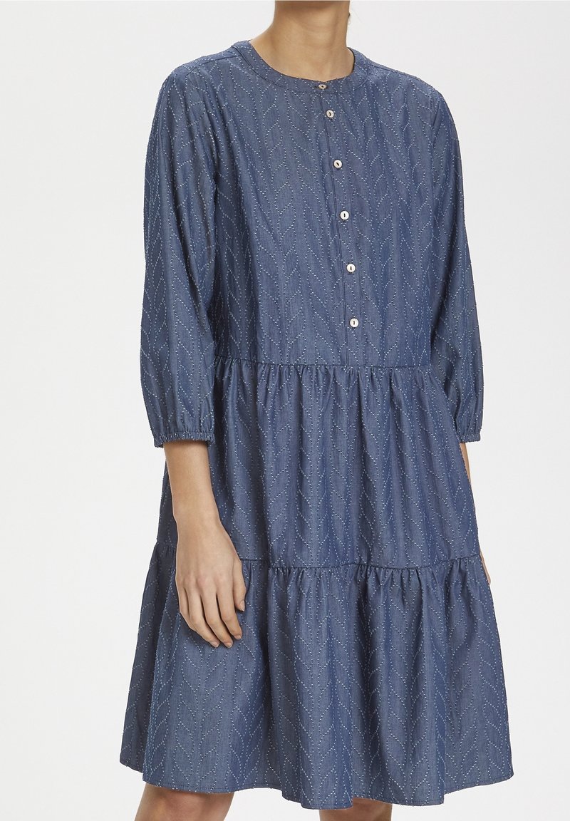 Modré šaty ke kolenům s texturovaným vzorem listů, dlouhé rukávy s elastickými manžetami a předním zapínáním na knoflíky na stojící osobě.