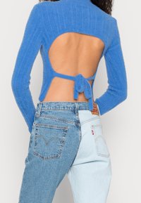 Maglione blu lavorato a maglia con schiena aperta e dettaglio con fiocco, abbinato a jeans in denim bicolore con una combinazione di blu chiaro e blu scuro.