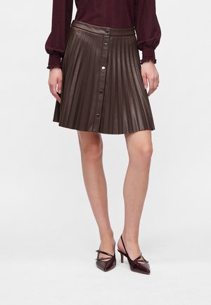 Femme portant une jupe en cuir plissée marron foncé avec des boutons sur le devant et des chaussures à talons aiguilles assorties et pointues.