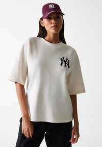 Cream ribbed oversized T-shirt with a round neckline and black NY logo. Paired with a maroon and white cap featuring embroidered logo.