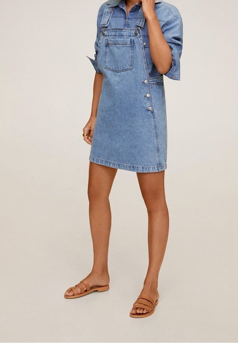 Robe salopette en jean bleu clair, avec une coupe structurée, une poche avant et des boutons latéraux décoratifs. Associée à des sandales marron à brides.