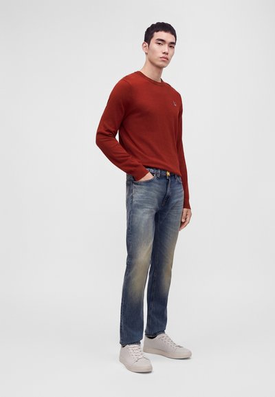 Jeune homme debout portant un pull à manches longues couleur rouille, un jean bleu délavé et des baskets blanches sur un fond gris clair uni.