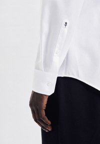 Camicia bianca con polsino abbottonato e ricamo scuro di una rosa sulla manica. Tessuto morbido e vestibilità sartoriale.