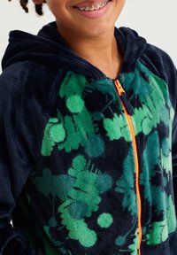 Sudadera con cremallera de forro polar azul marino con un patrón abstracto verde, cremallera naranja y una textura suave. Incluye un bolsillo tipo canguro en la parte delantera.