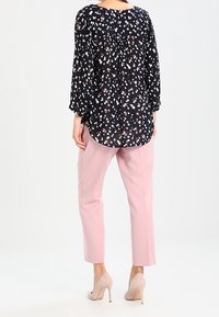 Schwarze Bluse mit weißem und rotem Blumenmuster, lässiger Schnitt und langen Ärmeln, kombiniert mit taillierten rosa Hosen und beigen Pumps.