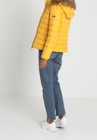 Żółta kurtka puchowa z kapturem wyłożonym futrem, zapinana na zamek z przodu, z kieszeniami po bokach. Połączona z niebieskimi dżinsami i białymi trampkami.