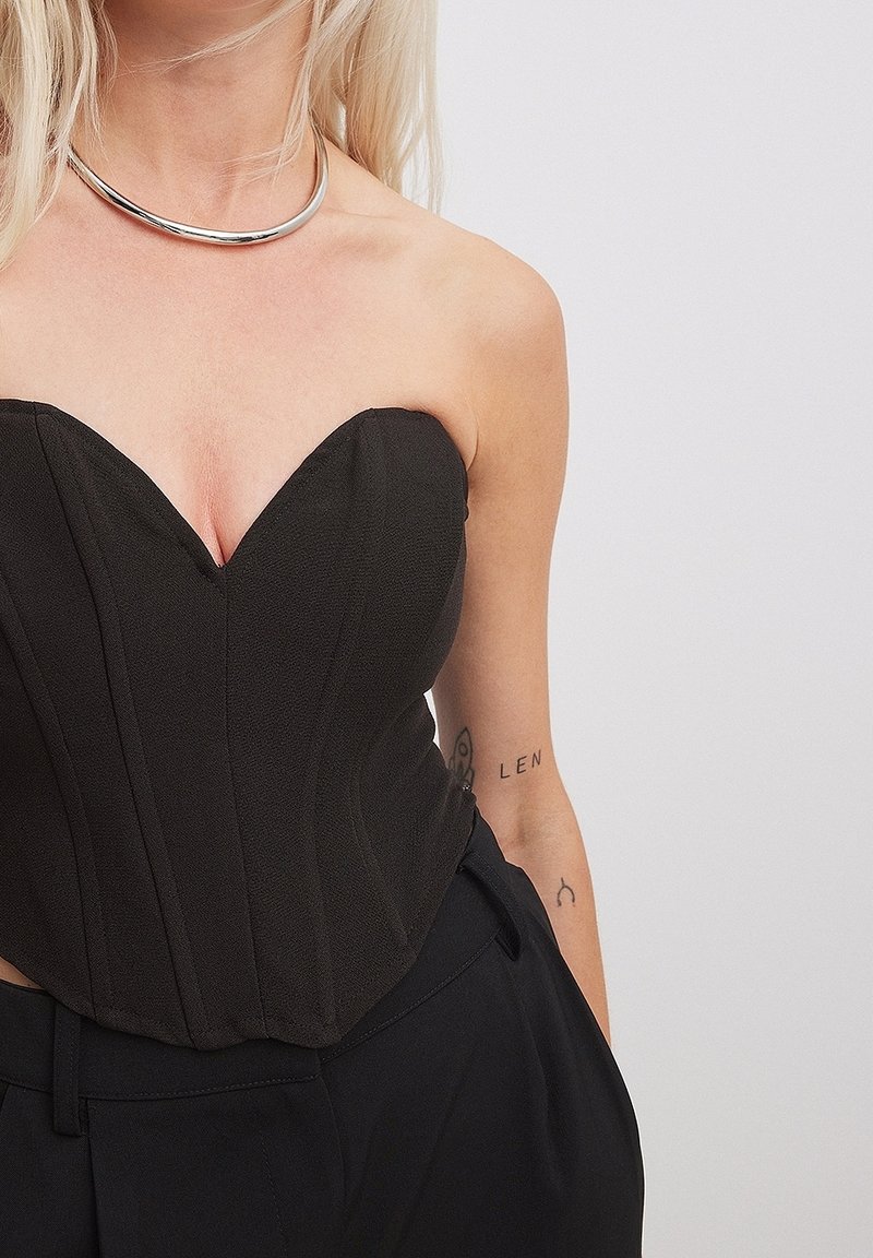 Femme blonde portant un corset noir sans bretelles, un pantalon noir et un collier argenté, avec de petits tatouages visibles sur son bras droit.