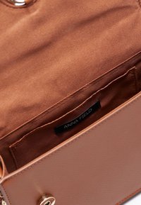 Borsa a tracolla di colore tan con finitura testurizzata, dotata di una chiusura in metallo dorato e foderata internamente di un morbido tessuto marrone, include un’etichetta con la scritta "ANNA FIELD."