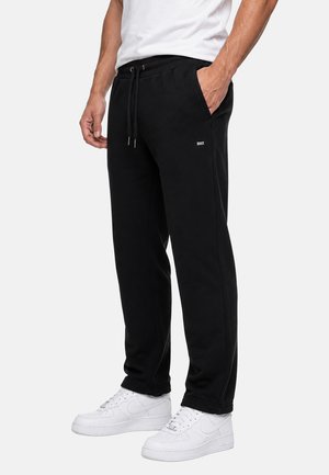 Uomo con pantaloni della tuta neri con coulisse e tasche laterali e sneakers bianche, in piedi con una mano in tasca davanti a uno sfondo semplice.