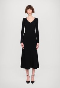 Abito lungo a maniche lunghe nero a coste con scollatura a V, corpetto aderente e gonna a ruota. Il modello indossa tacchi neri a punta e accessori semplici.