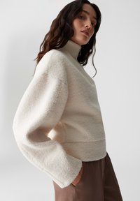 Cremefarbenes, strukturiertes Sweater mit hohem Kragen, langen Ärmeln und lockerer Passform. Mit seitlichen Schlitzen am Saum und einem weichen, fleece-ähnlichen Material.