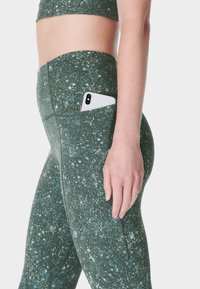 Vrouw in groen gespikkelde leggings met een smartphone die gedeeltelijk zichtbaar is in de zijzak, staand tegen een effen achtergrond.