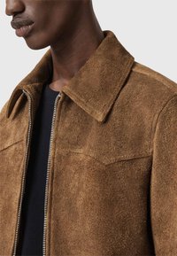 Ruskea mokkatakki, jossa on terävä kaulus, vetoketjusulku ja kaksi rintataskua. Pehmeä tekstuuri ja istuva malli.