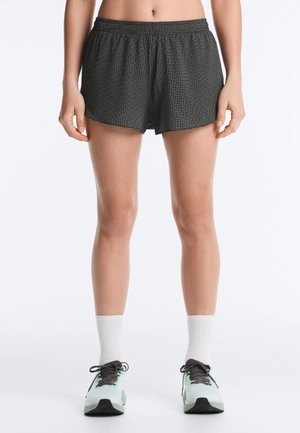 Persoon met zwart-witte geblokte shorts, witte sportsokken en witte en zwarte hardloopschoenen, staand tegen een witte achtergrond.