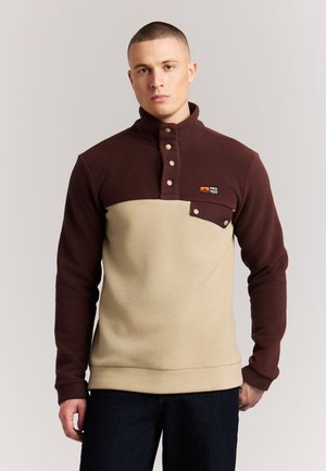 Jonge man met kort haar die een tweekleurige bruine en beige pullover van fleece draagt met een hoge kraag en een knoopzak op de borst, tegen een effen achtergrond.