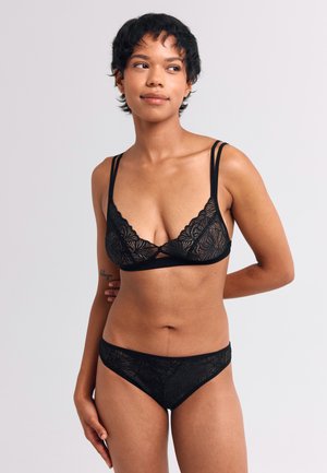 Zwarte kant bralette met een ingewikkeld bladpatroon, voorzien van twee dunne bandjes en een driehoekige vorm. Gepromoot met laag uitgesneden zwarte kanten slipjes.