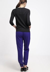 Zwarte nauwsluitende top met driekwart mouwen en een met kralen versierde halslijn, gecombineerd met losse paarse broek en zwarte platte schoenen.