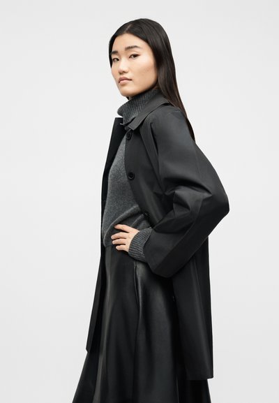 Γυναίκα με μακριά σκούρα μαλλιά, φοράει σκούρο γκρι πουλόβερ με ζιβάγκο, μαύρη δερμάτινη φούστα και μαύρο παλτό με κουμπιά, στέκεται πλαγίως με το χέρι στη μέση.