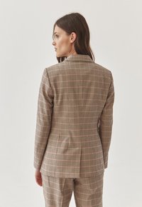 Blaser texturé à carreaux beige avec des touches de rouge et de vert, présentant une fente arrière unique et des revers structurés. Convient pour des tenues sur mesure.