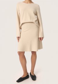 Beige strikket sweater og matchende flagrende nederdel, begge lavet af et blødt materiale. Sweateren har lange ærmer og en rund halsudskæring. Sorte flade sko.