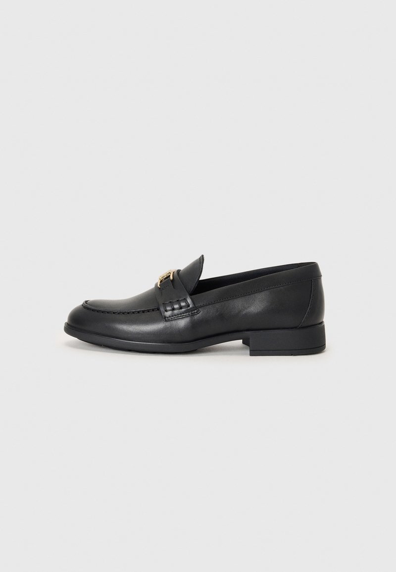 Mocassin en cuir noir à bout carré, talon bas et détail boucle dorée sur le dessus, sur fond clair uni.