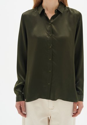 Kobieta ubrana w ciemnozieloną satynową koszulę na guziki z długimi rękawami oraz kremowe spodnie, pokazana od szyi do bioder.