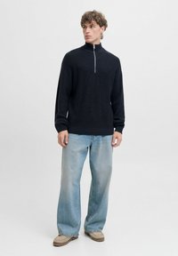 Czarny sweter z dzianiny na zamek błyskawiczny z wysokim kołnierzem, noszony na jasnoniebieskich spodniaczkach typu wide-leg i beżowych butach. Projekt charakteryzuje się prążkowaną teksturą.