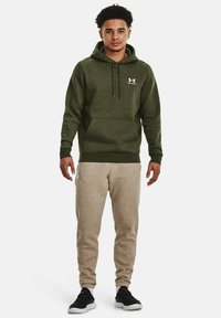 Olivengrøn hættetrøje med kengurulomme og snoretræk i hætten, parret med lys beige sweatpants og sorte sportssko.