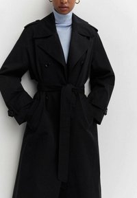 Černý trench coat s dvouřadým zapínáním, velkými klopami, páskem v pase a bočními kapsami. Materiál vypadá hladce; zahrnuje detaily s knoflíky.