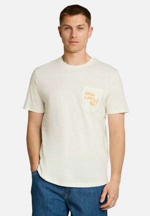 Mann trägt ein hellcremefarbenes T-Shirt mit einer Tasche, auf der der Text "Fresh & Juicy" und ein Orangenscheiben-Design abgebildet sind, kombiniert mit blauen Jeans.