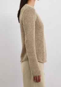 Femme portant un pull beige en maille épaisse à manches longues, vue de profil sur un fond uni.