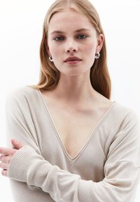 Suéter de punto beige con un profundo escote en V, corte holgado y puños acanalados. La modelo tiene el cabello largo y lleva pendientes circulares de plata.