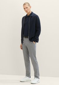 Marineblauwe geribbelde cardigan over een marineblauw polo, gecombineerd met lichtgrijze broeken en witte sneakers. Eenvoudig ontwerp met ritsluiting en subtiele textuur.
