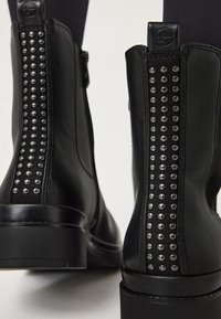 Bottines en cuir noir avec des rangées de clous argentés le long de la couture arrière et des petits talons blocs, portées avec des collants noirs.