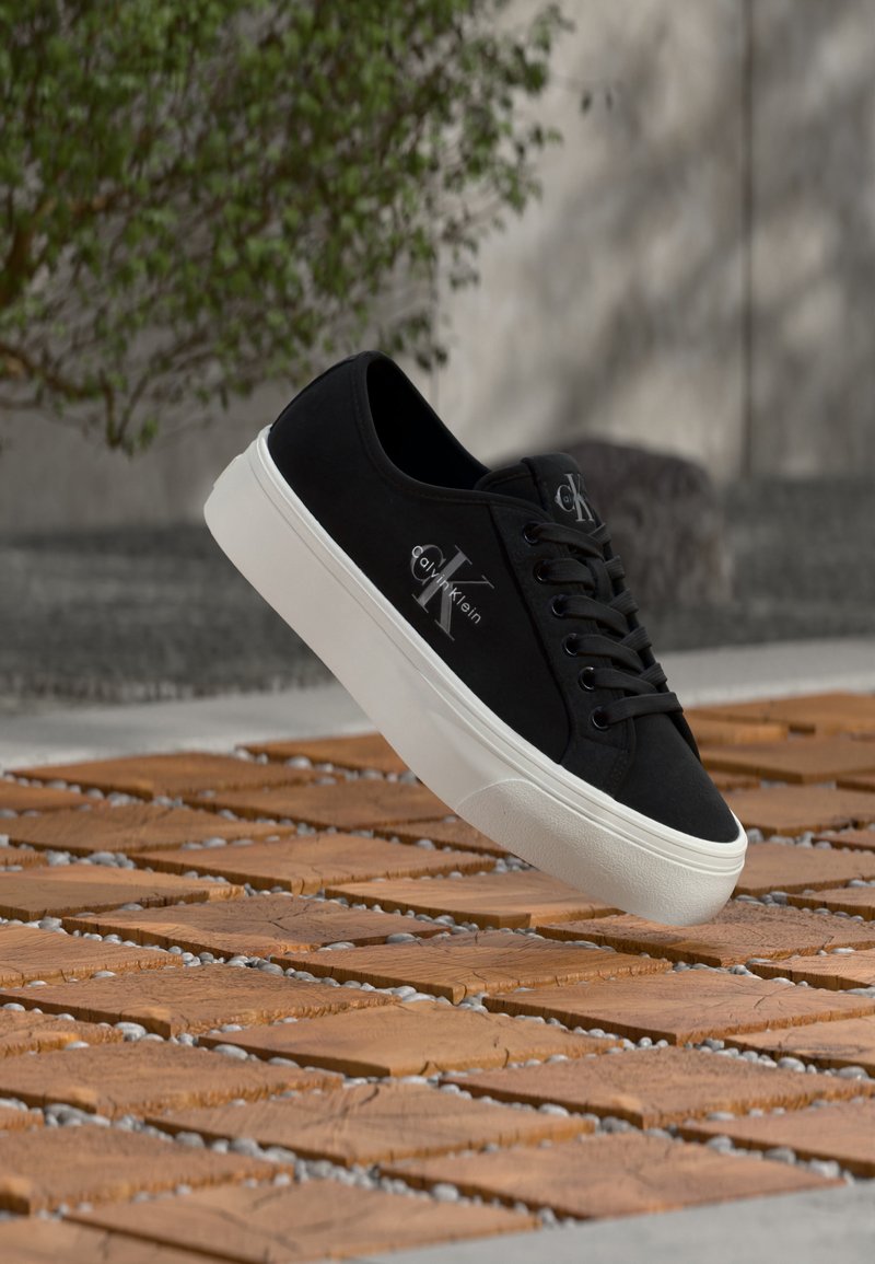 Baskets basses noires avec semelle blanche et logo "Calvin Klein", flottant au-dessus d'une surface en briques texturée avec de la verdure et un mur en béton en arrière-plan.