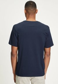 Man med kort hår som bär en enkel marinblå t-shirt med kort ärm och ljusbeiga byxor, vänd bort från kameran mot en vit bakgrund.